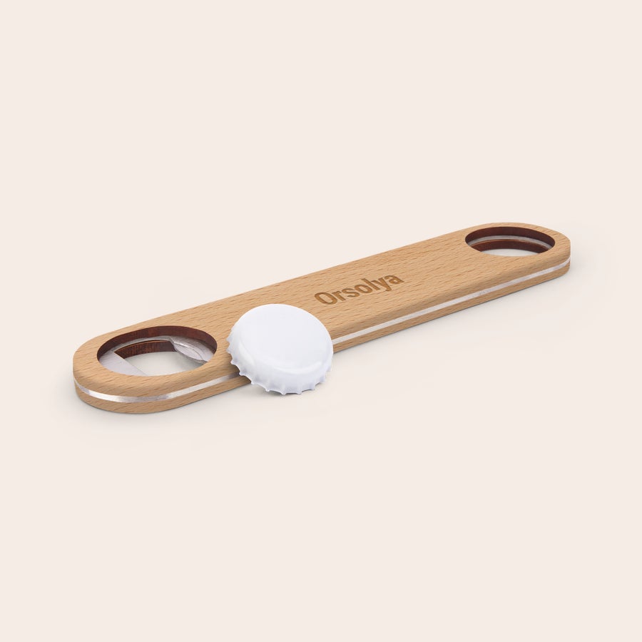 Gravírozott egyedi fa sörnyitó Személyre szabott fából készült sörnyitó Orsolya névvel gravírozva, mellette egy fehér üvegkupak fekszik