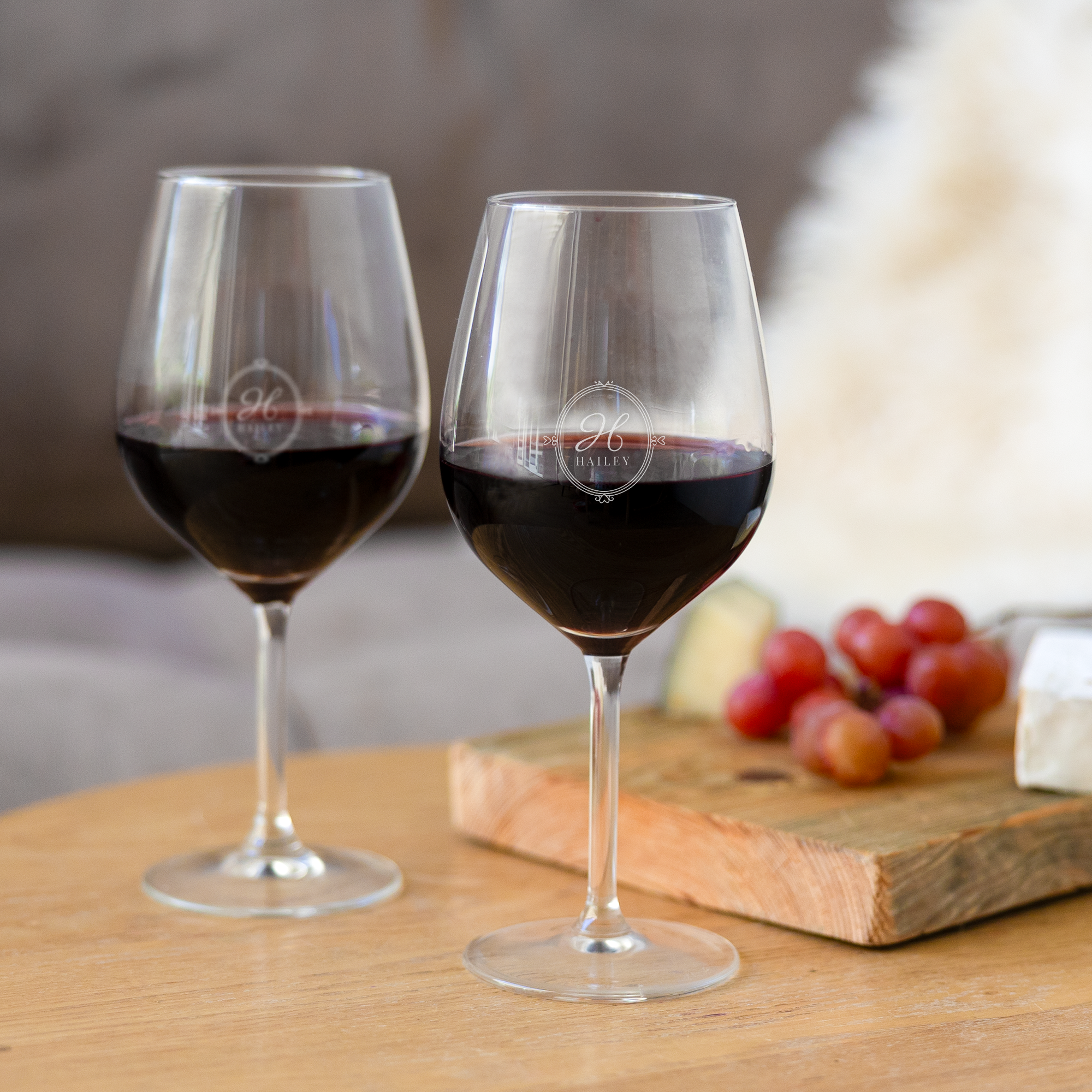 Due calici da vino rosso personalizzati, incisi con iniziale H e nome Hailey, su un tavolo con formaggio e uva.