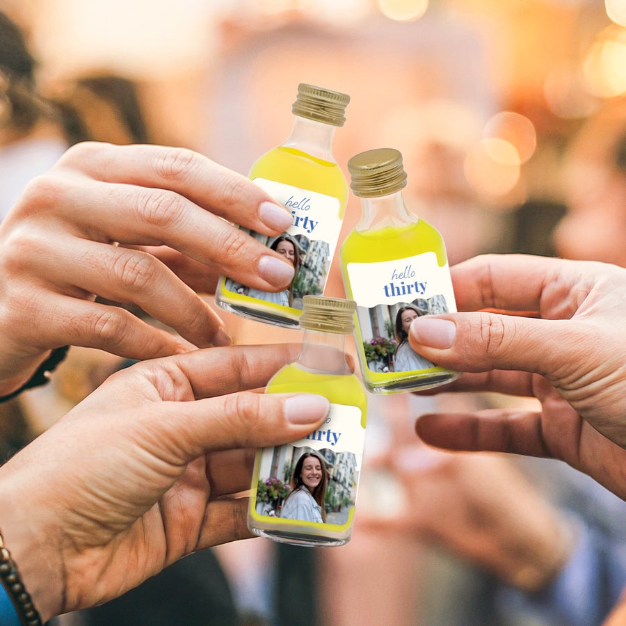 Personalised mini alcohol bottles Three hands holding up mini limoncello bottles with custom photo labels and "hello thirty" text to create a unique Mini Limoncello collection.