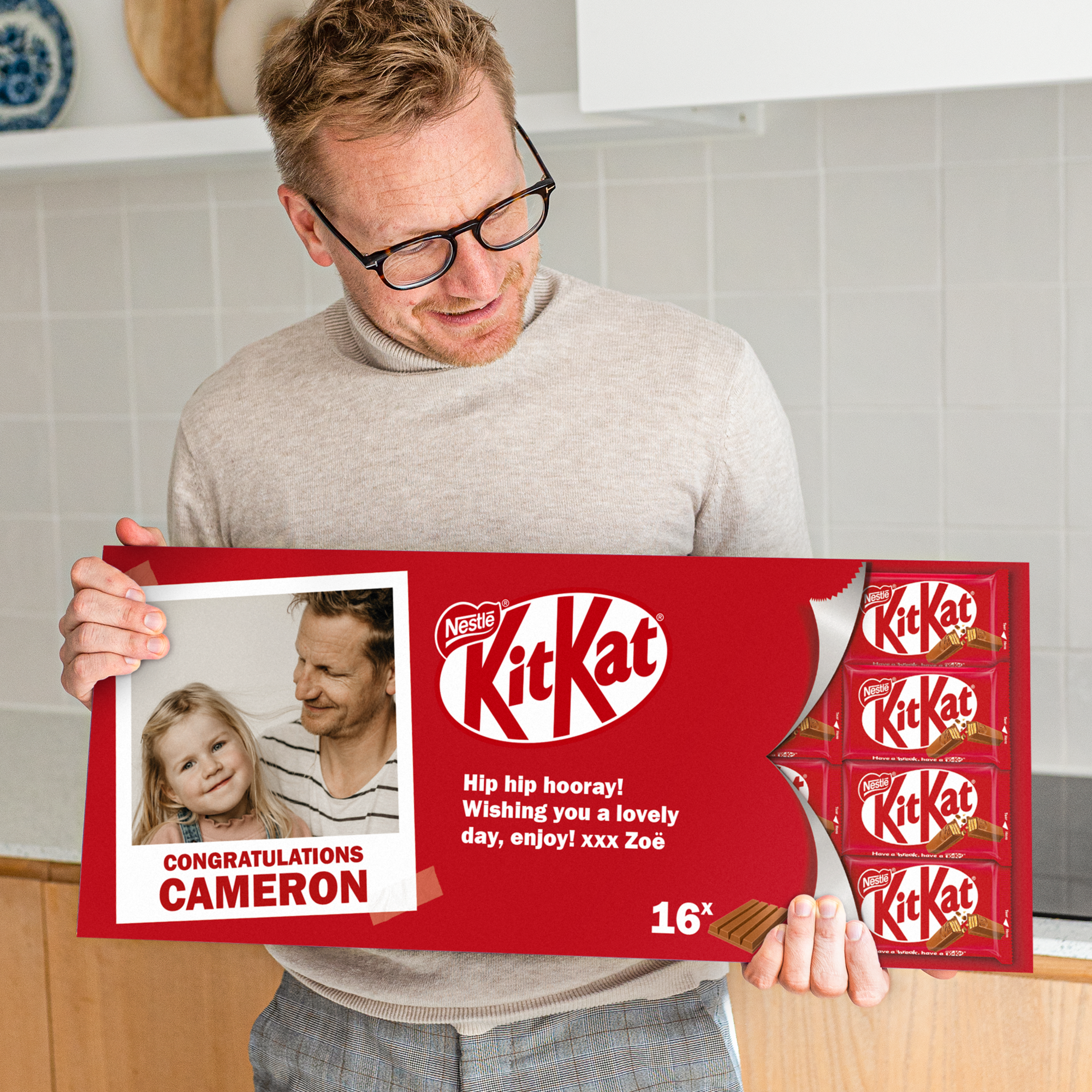 A man holding a personalized giant KitKat chocolate bar printed with a photo of a man and child, text "CONGRATULATIONS CAMERON", and a message "Hip hip hooray! Wishing you a lovely day, enjoy! xxx Zoë".