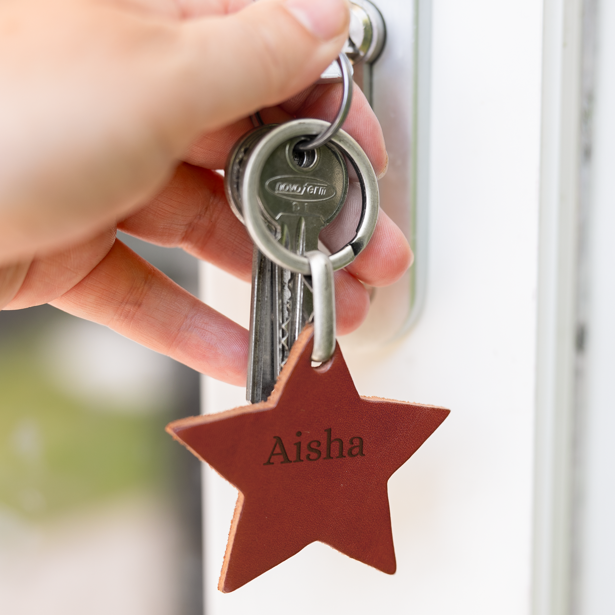 Hand die een stervormige leren sleutelhanger met naam 'Aisha' en sleutels vasthoudt, gegraveerd met persoonlijke tekst.