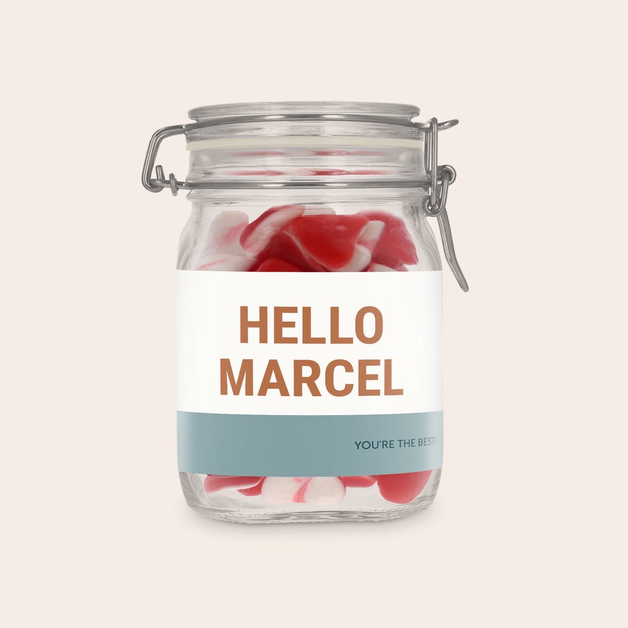 Személyre szabott édességes üveg Személyre szabott édes tégely szív alakú cukorkákkal, "HELLO MARCEL" felirattal, egyedi finomság matricával az üvegen.