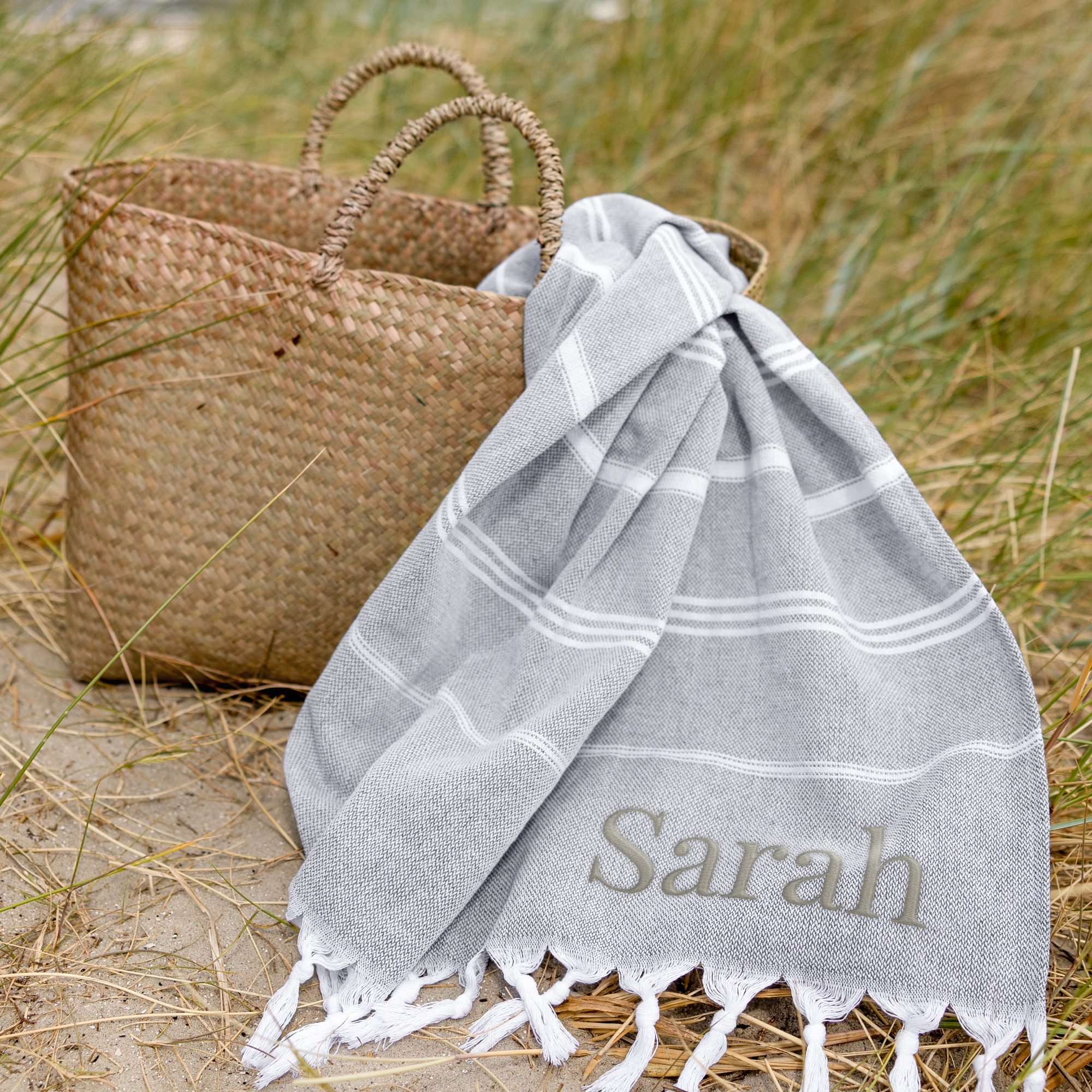 Toalla hammam personalizada gris con nombre bordado Sarah, junto a una cesta de mimbre en la arena.