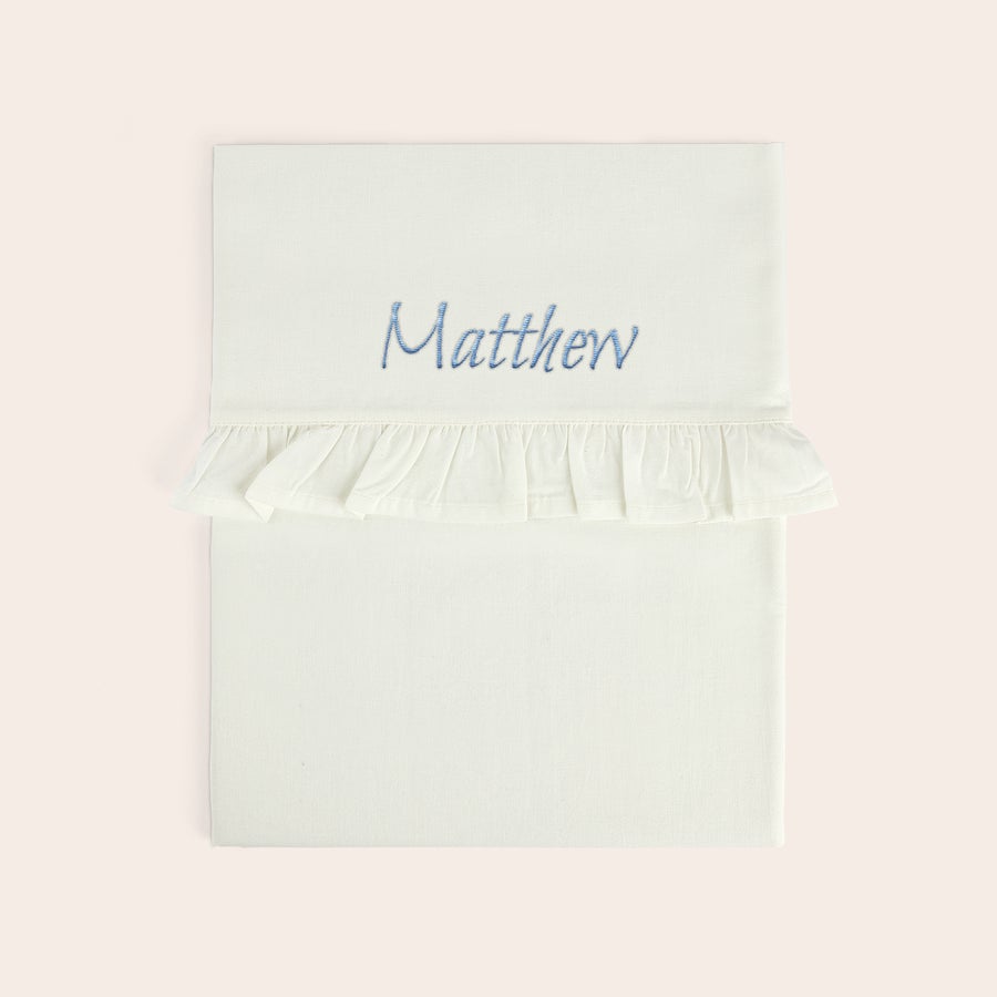Sábana para cuna bordada - Borde con volantes Sábana de moisés blanca con volante, bordada con el nombre "Matthew" en azul.
