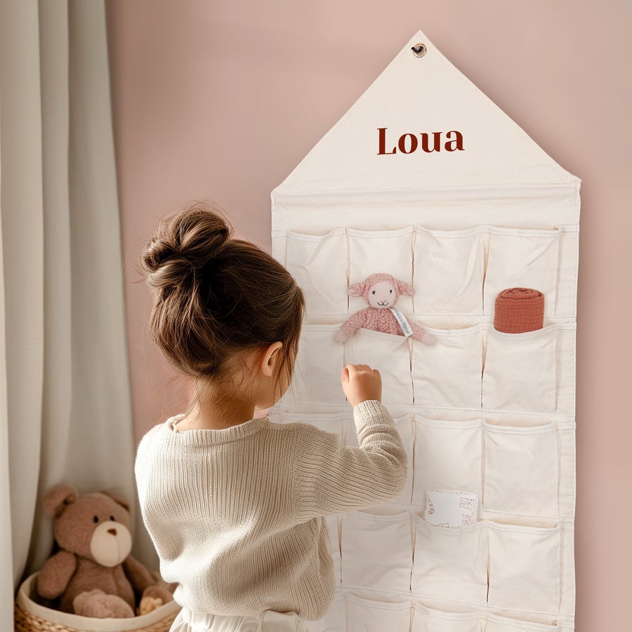 Organizator de buzunar din material textil Organizator personalizat de buzunar din material textil bej cu numele Loua imprimat, umplut cu jucarii si o fetita care il aranjeaza