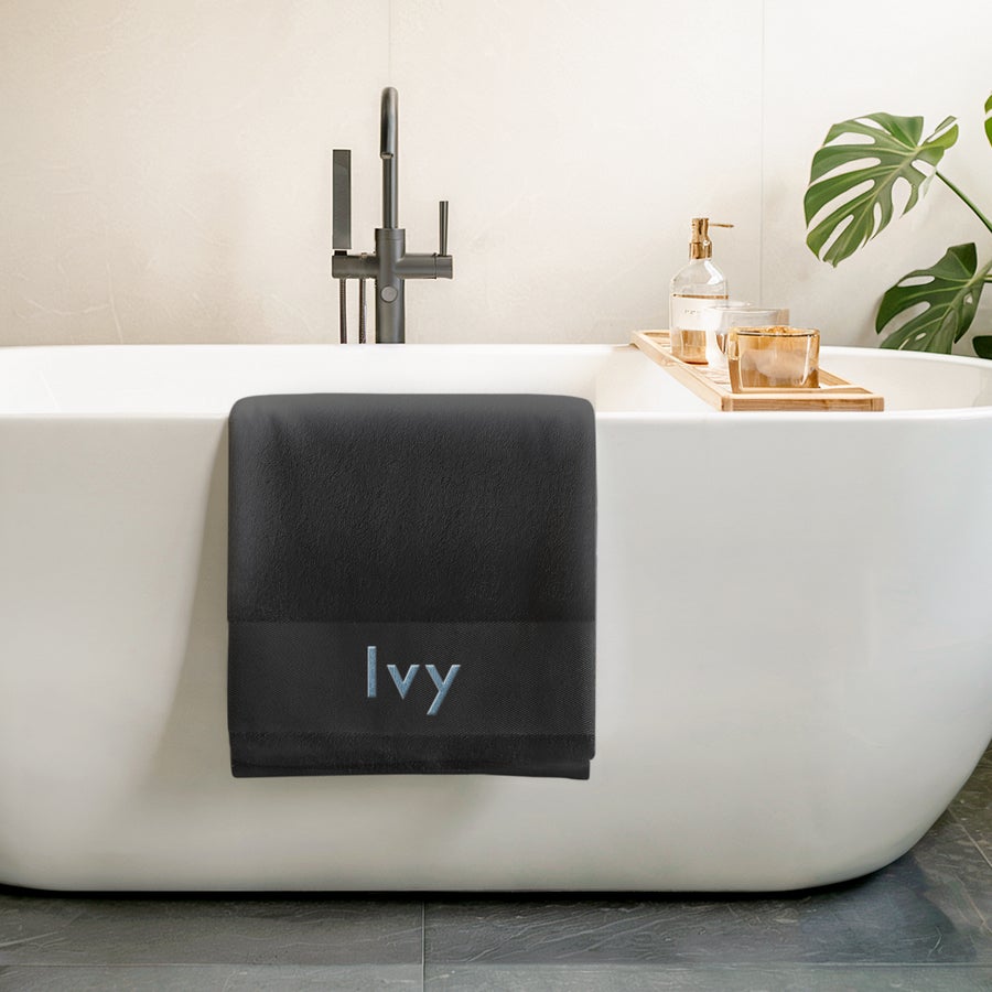 Asciugamano da bagno con testo Asciugamano nero personalizzato ricamato con il nome Ivy in celeste, appoggiato a una vasca da bagno. Crea qualcosa di unico.