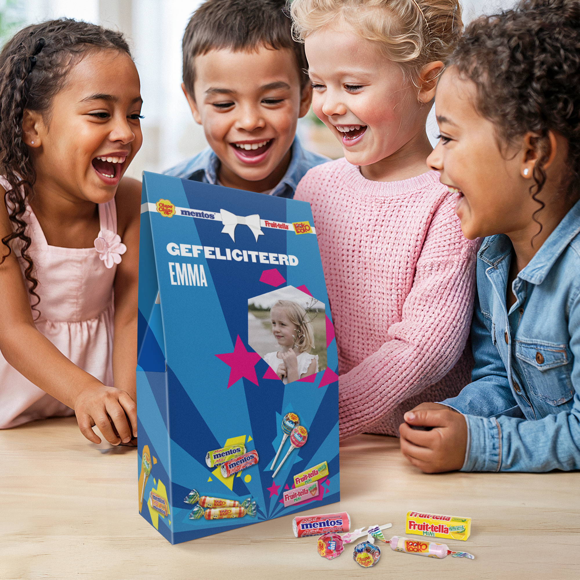 Vier blije kinderen kijken naar een gepersonaliseerd Fruit-tella cadeau, bedrukt met de tekst "Gefeliciteerd Emma" en een foto van een meisje.