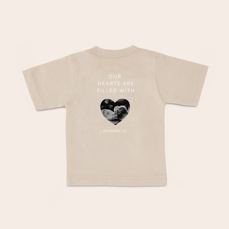 T shirt bébé personnalisé T-shirt bébé beige personnalisé avec échographie en forme de cœur, texte "Our hearts are filled with" et date imprimés.