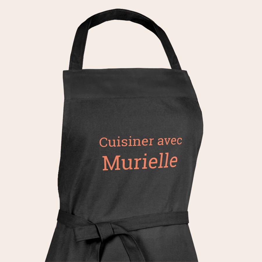Tablier personnalisé Offrez un tablier de cuisine noir personnalisé, imprimé avec le texte "Cuisiner avec Murielle" en orange