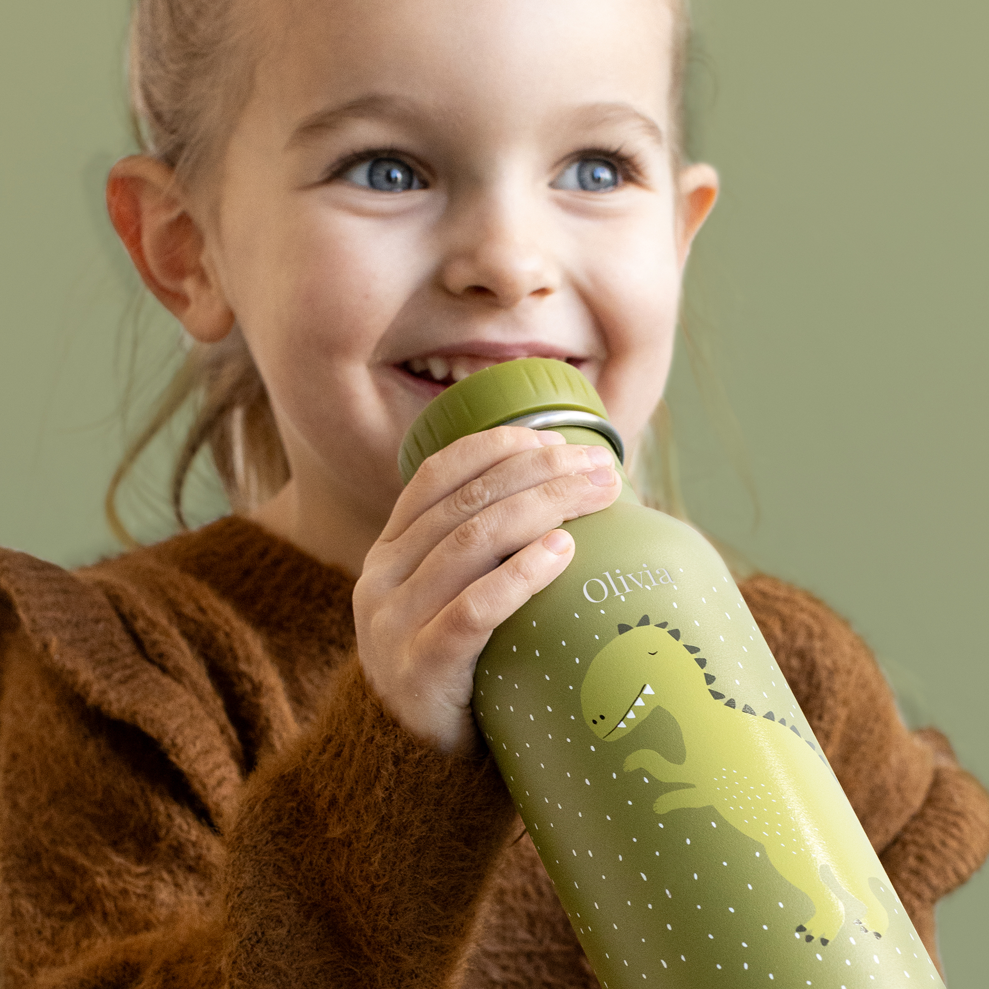 Bambina che tiene una bottiglia d'acqua a forma di dinosauro Trixie personalizzata, con il nome Olivia inciso al laser, sorride e guarda in alto.