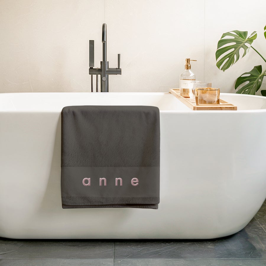 Toalha de banho com texto Toalha de banho antracite personalizada com o nome Anne bordado a rosa, pendurada numa banheira branca. Cria algo único com uma toalha de banho antracite personalizada.