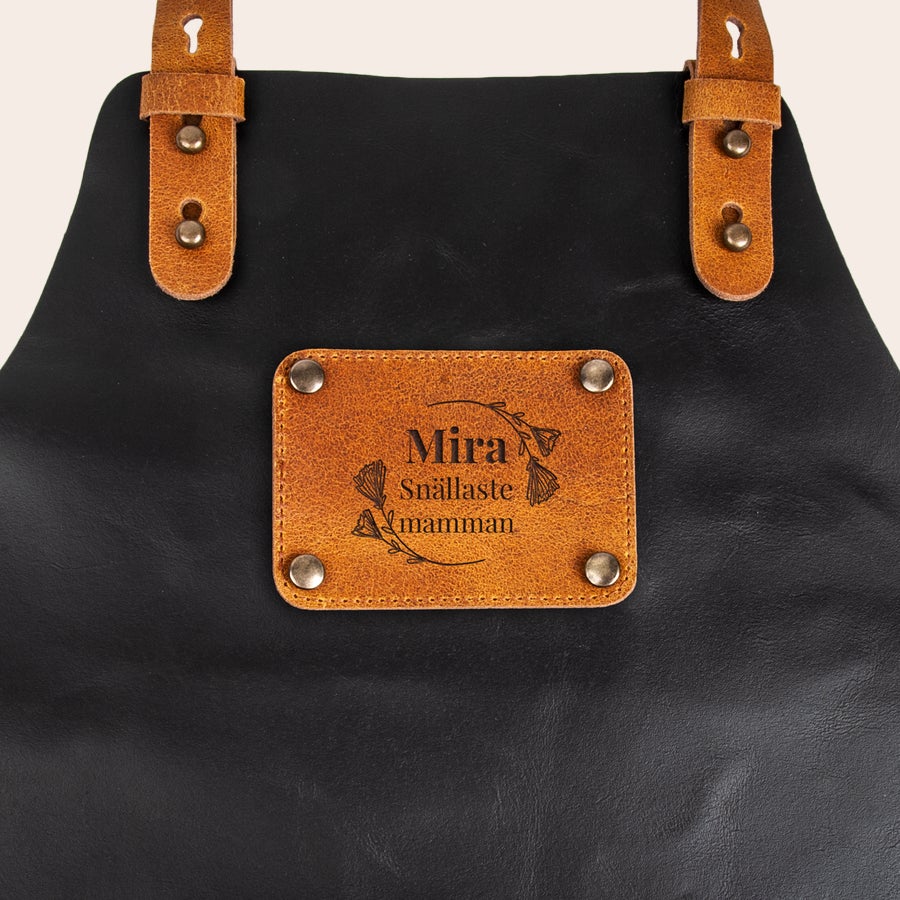 Läderförkläde med namn Svart läderförkläde med ljusbrun läderlapp, graverad med Mira Snällaste mamman. Visa vem som är kock med detta fantastiska läderförkläde med namn!