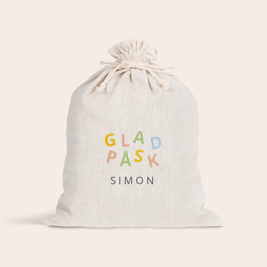 Presentpåse med dragsko - påsk Vit presentpåse med dragsko, tryckt med texten "GLAD PÅSK" i olika färger och namnet "SIMON", för att göra påsken speciell.