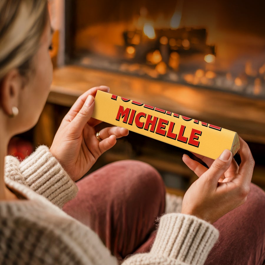 Toblerone med navn Personaliseret Toblerone med navn og billede, med navnet Michelle trykt på chokoladeindpakningen, holdt af en person foran en pejs.