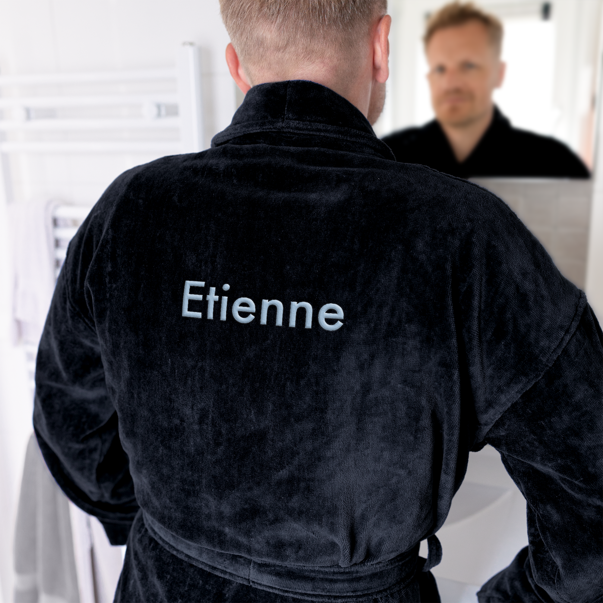 Peignoir noir doux brodé du prénom Etienne, vu de dos dans une salle de bain, reflet de l'homme dans le miroir.
