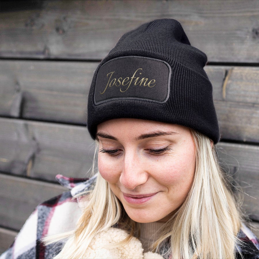 Hue med navn - Broderet Ung kvinde med sort beanie med "Josefine" broderet i guld på en sort lap, som holder ørerne varme.