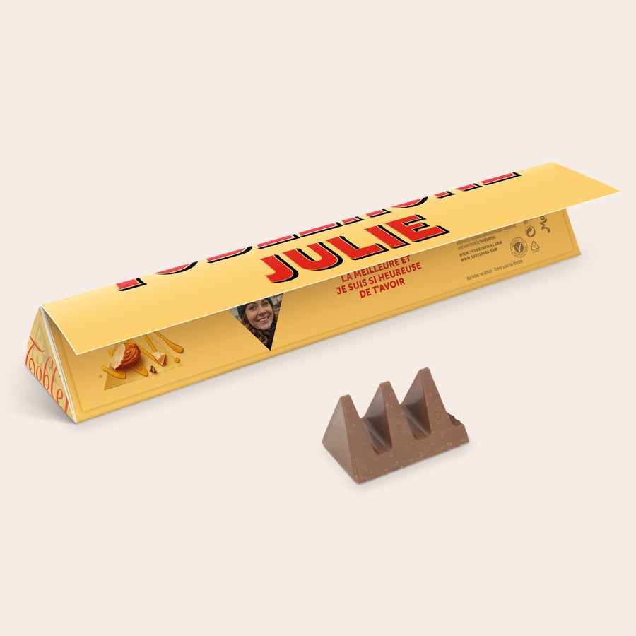 Toblerone personnalisé Toblerone personnalisé avec prénom et photo - S, imprimé avec le nom Julie et un message sur son emballage jaune, à côté d'une barre de chocolat.