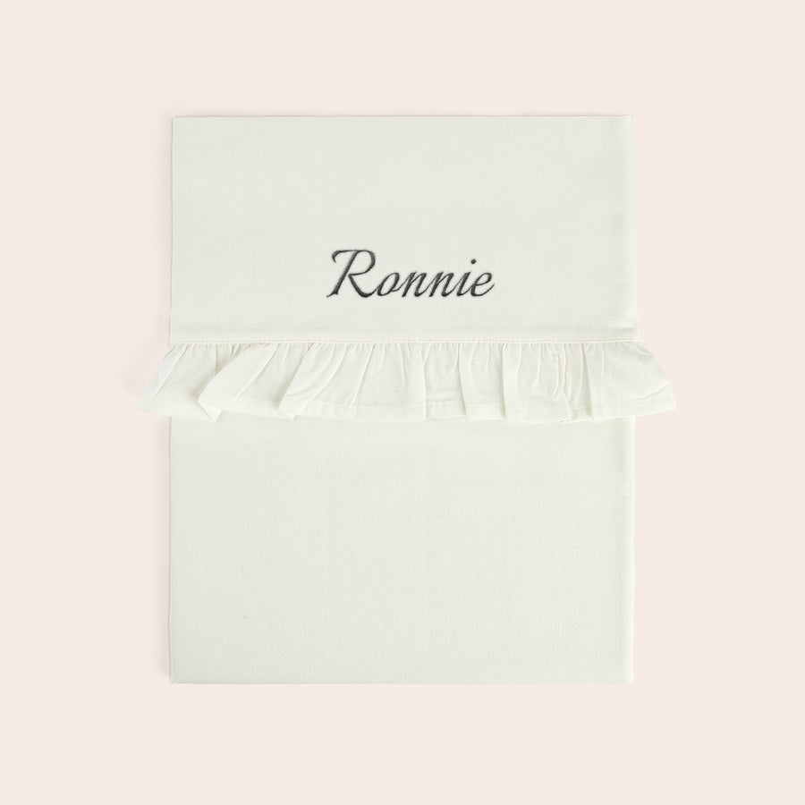 Sábana para cuna bordada - Borde con volantes Sábana de moisés blanca con el nombre Ronnie bordado en gris, con un borde de volantes decorativo.