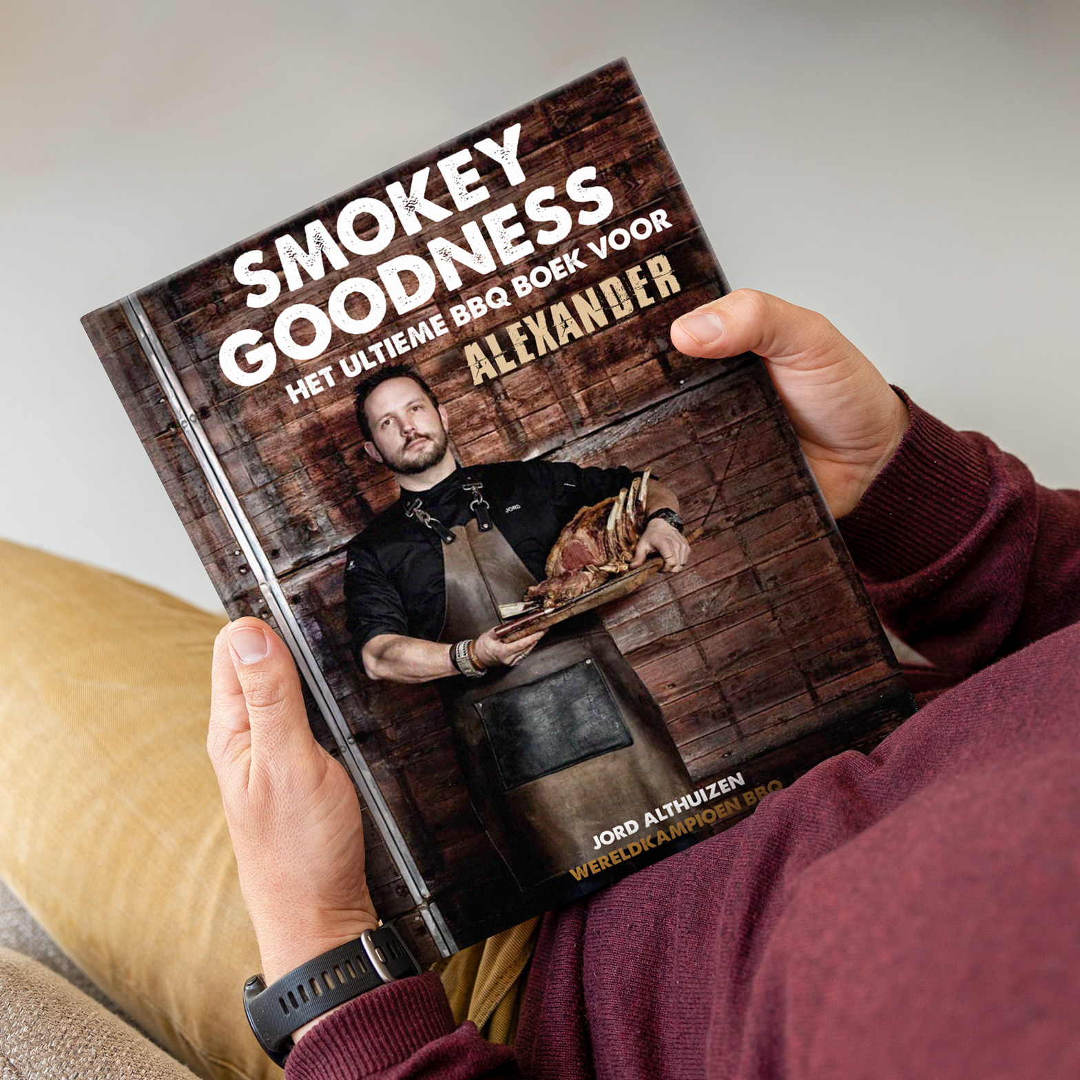 Persoonlijk Smokey Goodness BBQ boek met eigen naam Alexander op de kaft, vastgehouden door iemand in een bordeauxrode trui.