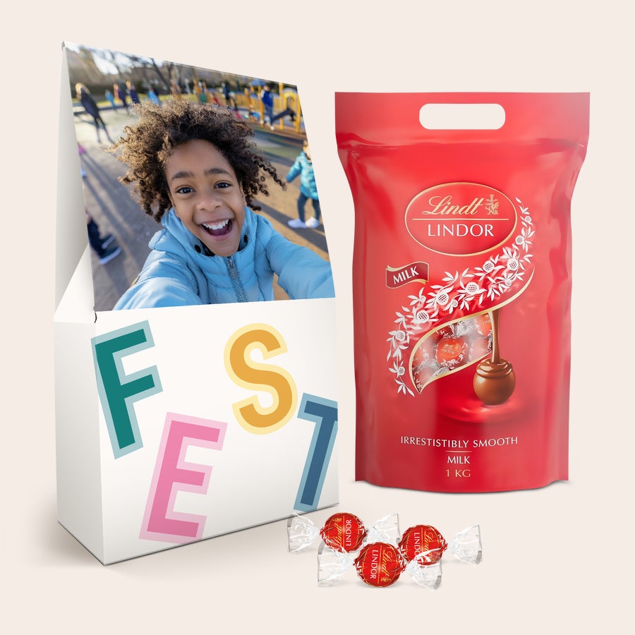Lindt 1kg Schokolade Geschenkbox Geschenkbox mit einem lachenden Kind auf dem Deckel, beschriftet mit "FEST", und einem Kilo Lindt Lindor-Milchschokolade zum Überraschen.