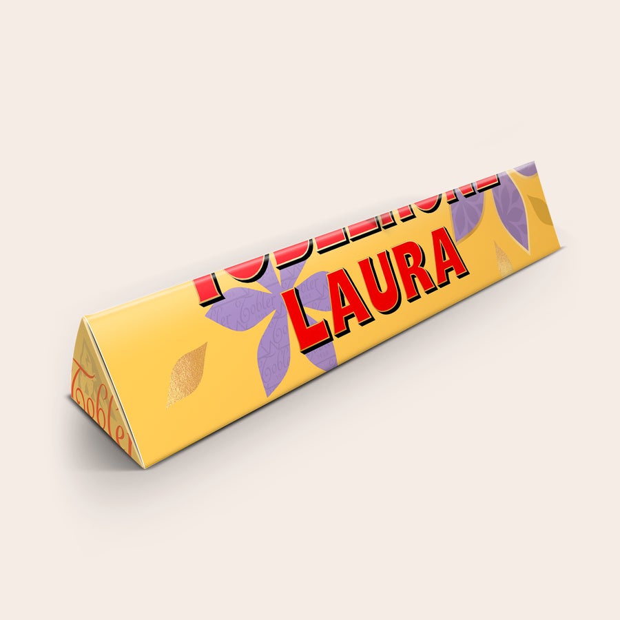 Toblerone Best Mom Personalisieren Personalisierte Toblerone Schokolade mit dem Namen Laura und lila Blüten gedruckt. Überraschen Sie Ihre Mama mit einer personalisierten Tafel Toblerone-Schokolade.