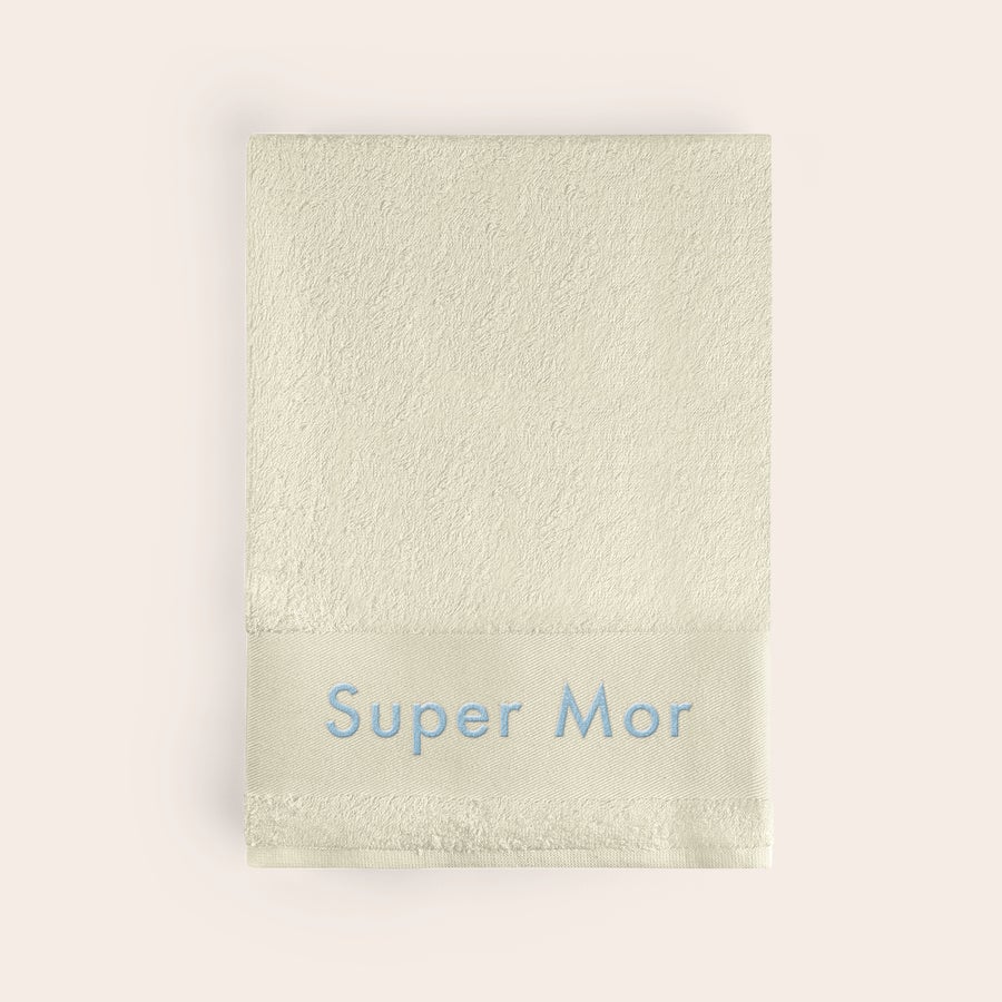 Håndklæde med tekst Beige håndklæde med broderet teksten "Super Mor" i lyseblå, gør hvert bad til noget særligt med et håndklæde i beige med broderi