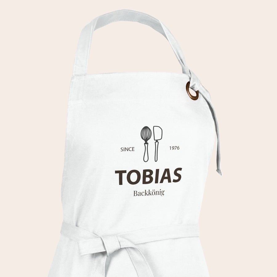Schürze bedrucken Weiße Schürze mit aufgedrucktem Design: Schneebesen, Pfannenwender, "Since 1976", dem Namen "Tobias" und "Backkönig"