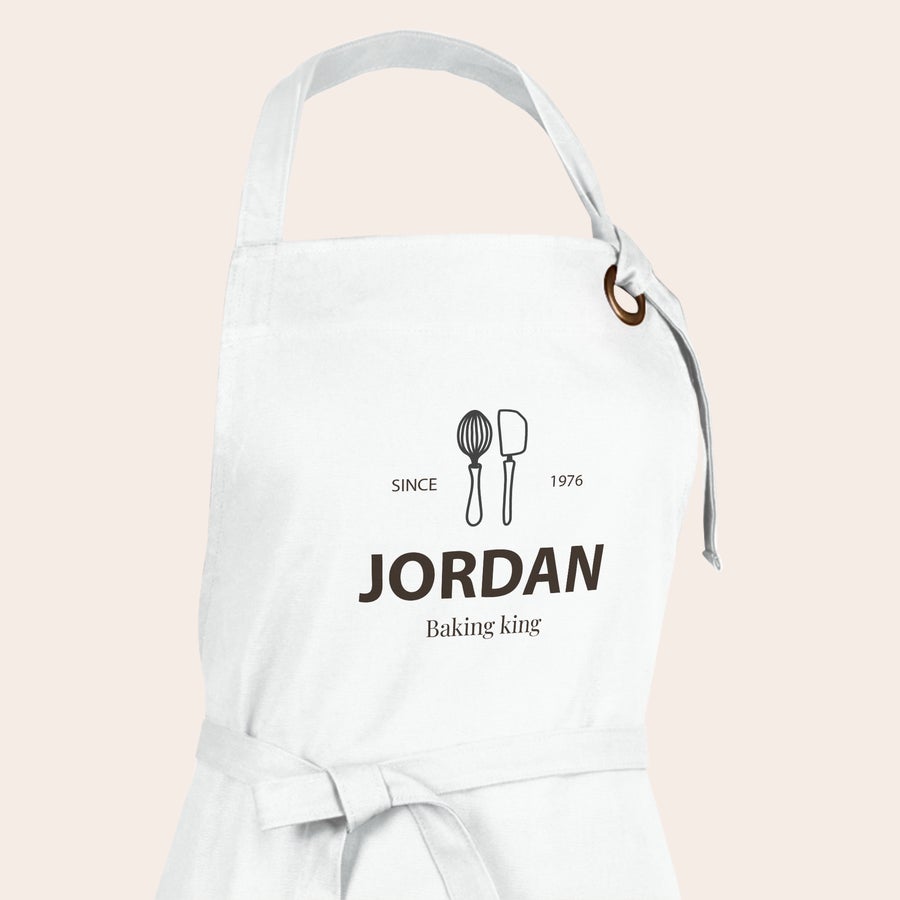 Forkle Hvitt kjøkkenforkle med trykt design av visp og slikkepott, samt tekst "JORDAN Baking king" og "SINCE 1976"