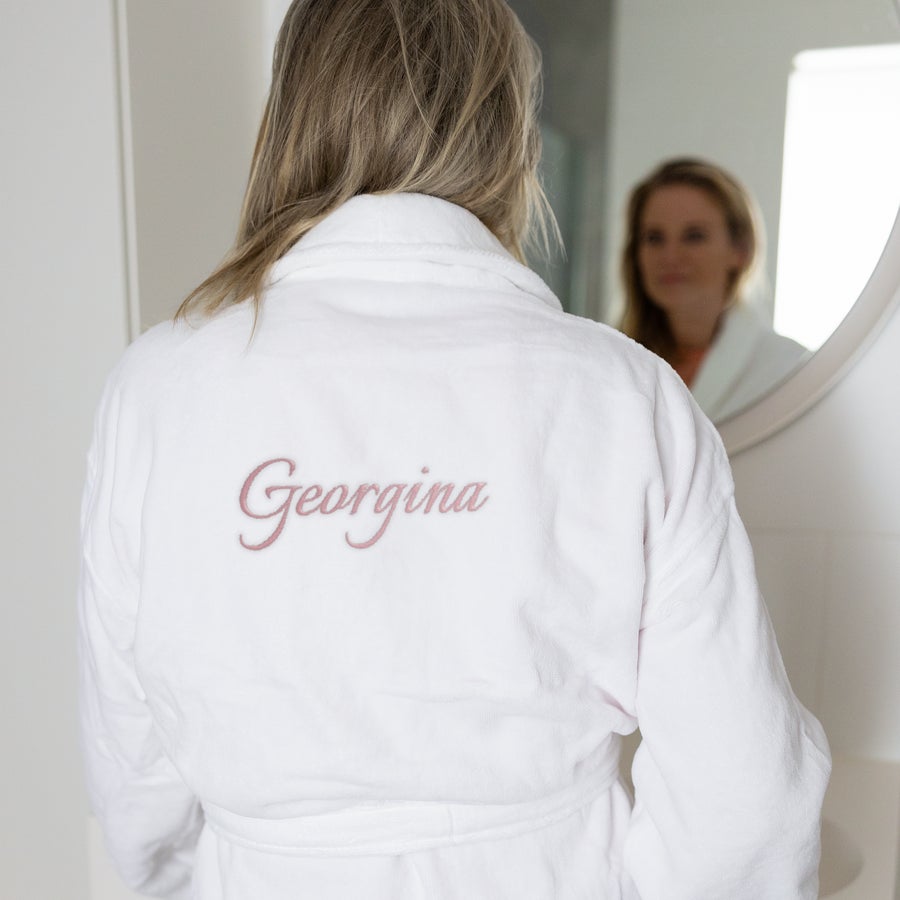 Kopalni plašč - ženske Beli kopalni plašč z vezenim imenom Georgina in žensko, ki se gleda v ogledalo, podarite mehak kopalni plašč