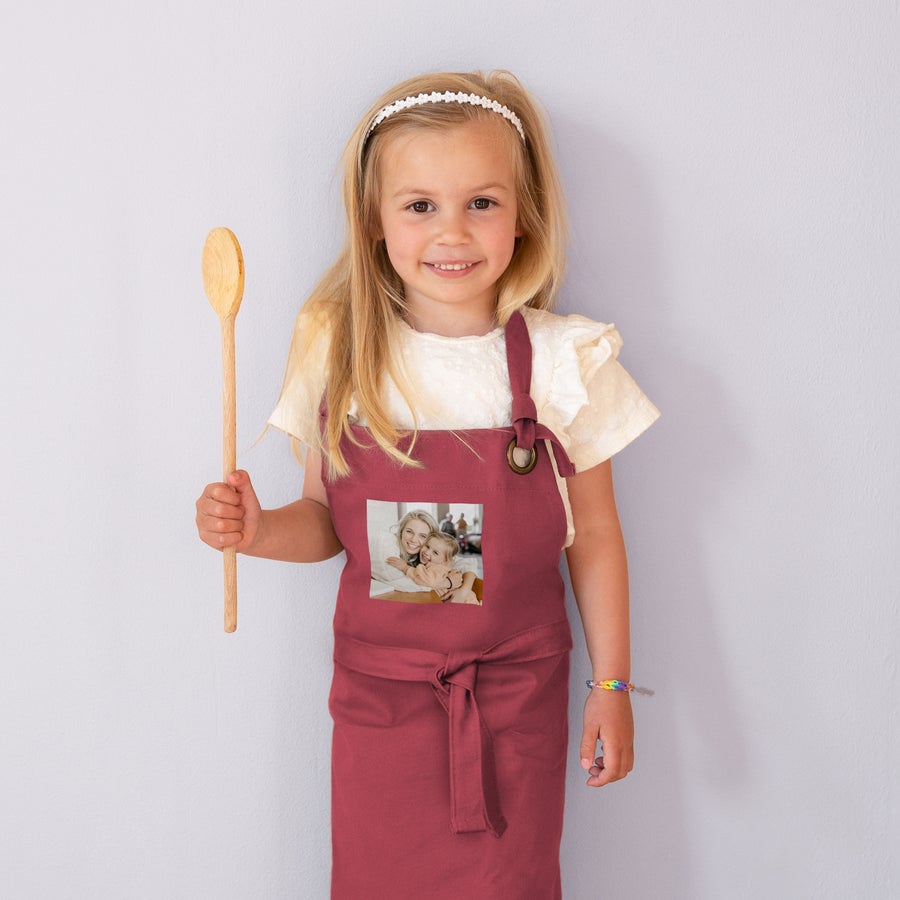 Delantal infantil personalizado Niña con un delantal personalizado color granate impreso con una foto de mamá y bebé, sosteniendo una cuchara de madera.