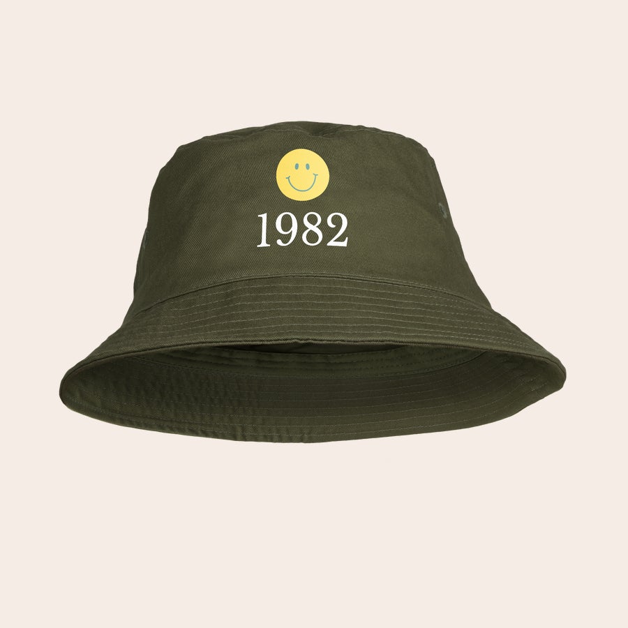 Bucket hat bedrukken Donkergroene zonnehoed bedrukt met een gele smiley en het jaartal 1982, om de zonnehoed met naam of foto te personaliseren.