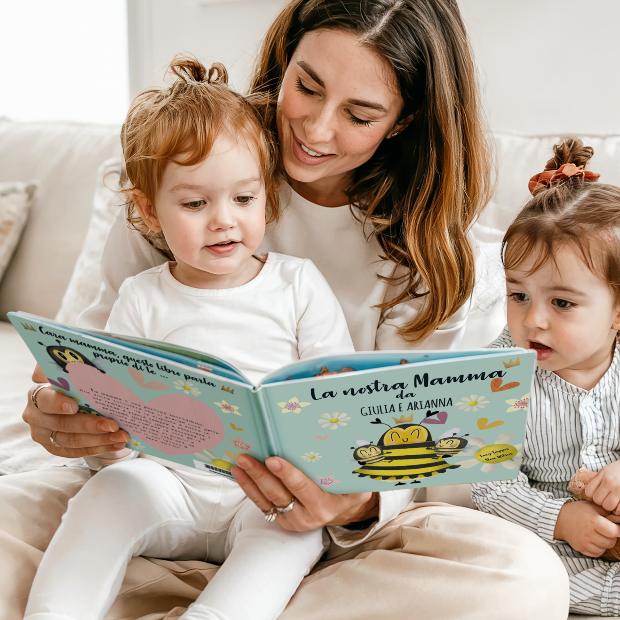 Una mamma legge il libro personalizzato "La nostra Mamma" con le figlie. La copertina è personalizzata con i nomi Giulia e Arianna e un'illustrazione di api. Crea una storia bellissima con te e la tua mamma come protagonisti!
