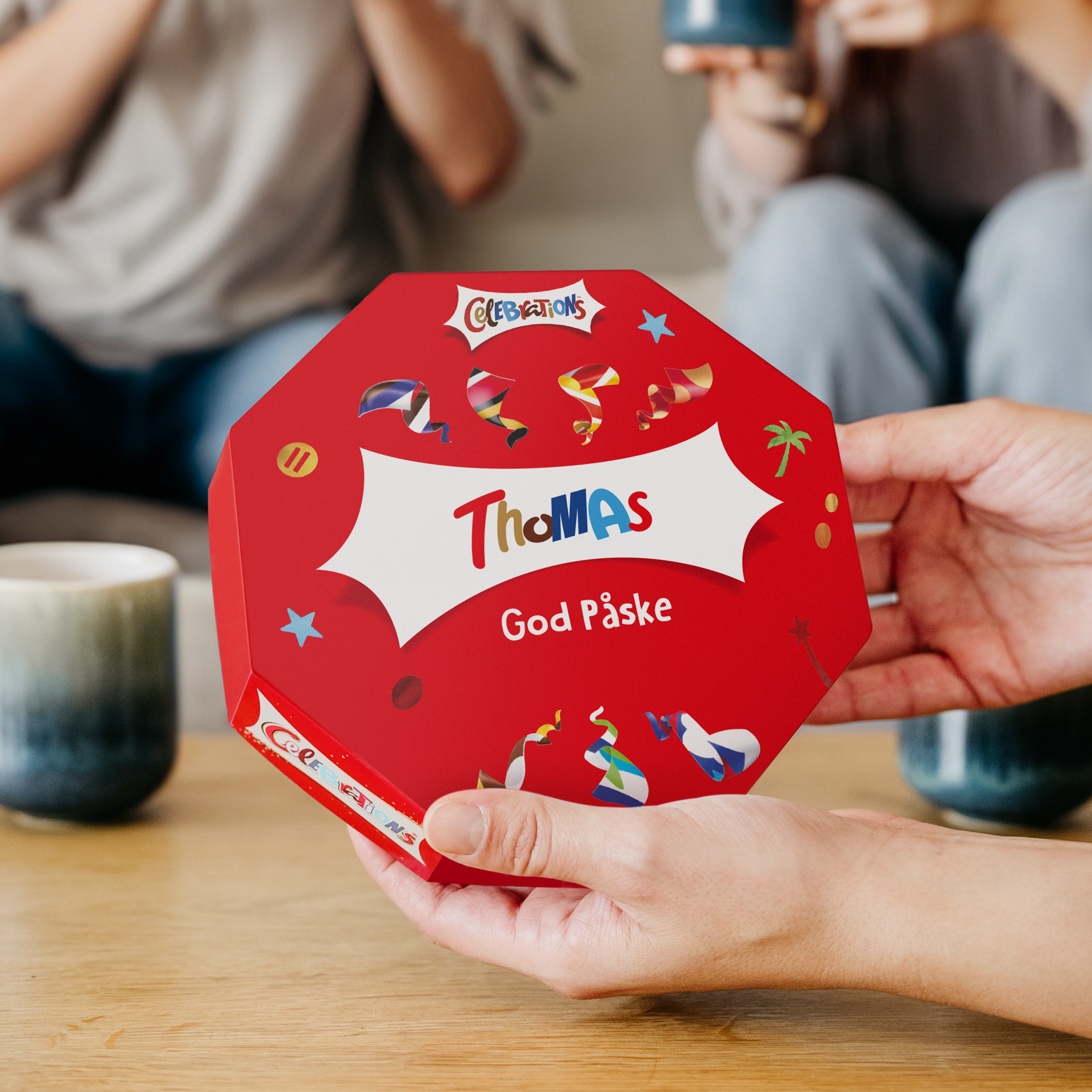 Rød Celebrations-eske med navn og God Påske tekst, trykket med personlig hilsen, holdes i hender.