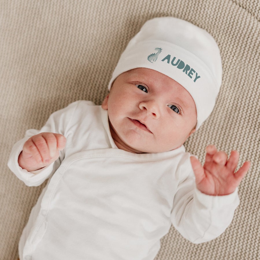 Bonnet naissance personnalisé Bébé porte un bonnet de naissance blanc imprimé avec un dinosaure et le prénom Audrey pour un cadeau de naissance