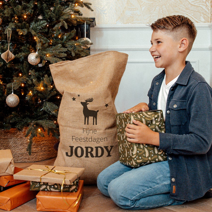 Jute kerstzak met naam bedrukt Jonge jongen met gepersonaliseerde jute kerstzak met naam Jordy en opdruk van rendier, naast kerstboom met kerstcadeaus.